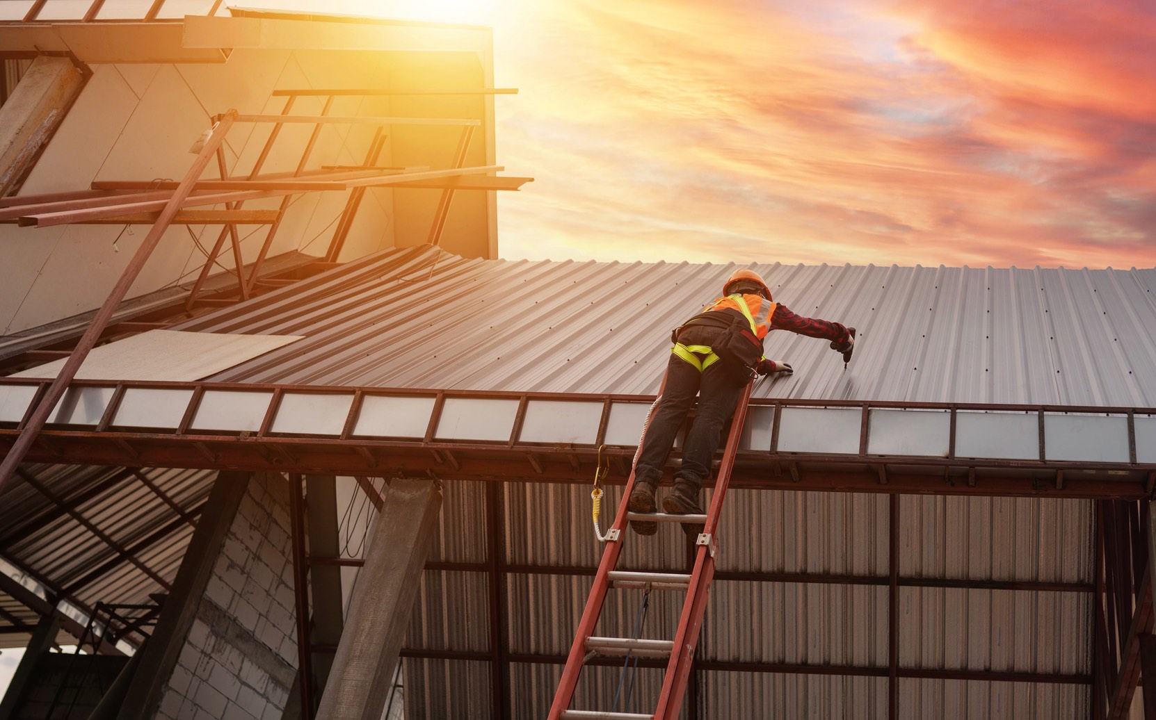 Dachdecker mit Schutzhelm auf einer Leiter montiert ein Metalldach, dahinter Abendhimmel im Sonnenuntergang