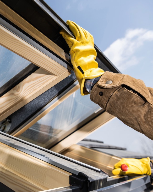 Hände in gelben Arbeitshandschuhen setzen ein Dachfenster mit Holzrahmen ein