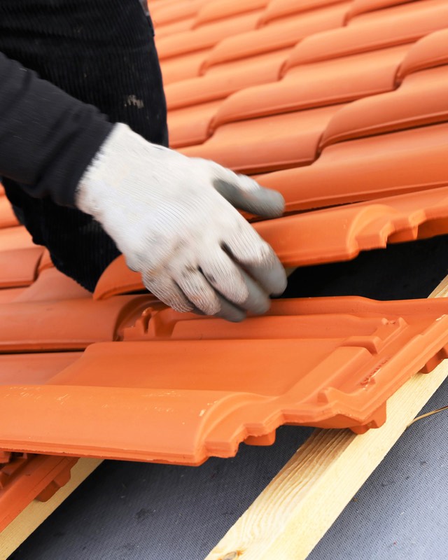 Hände in Arbeitshandschuhen verlegen rote Tondachziegel auf neuer Dachlattung
