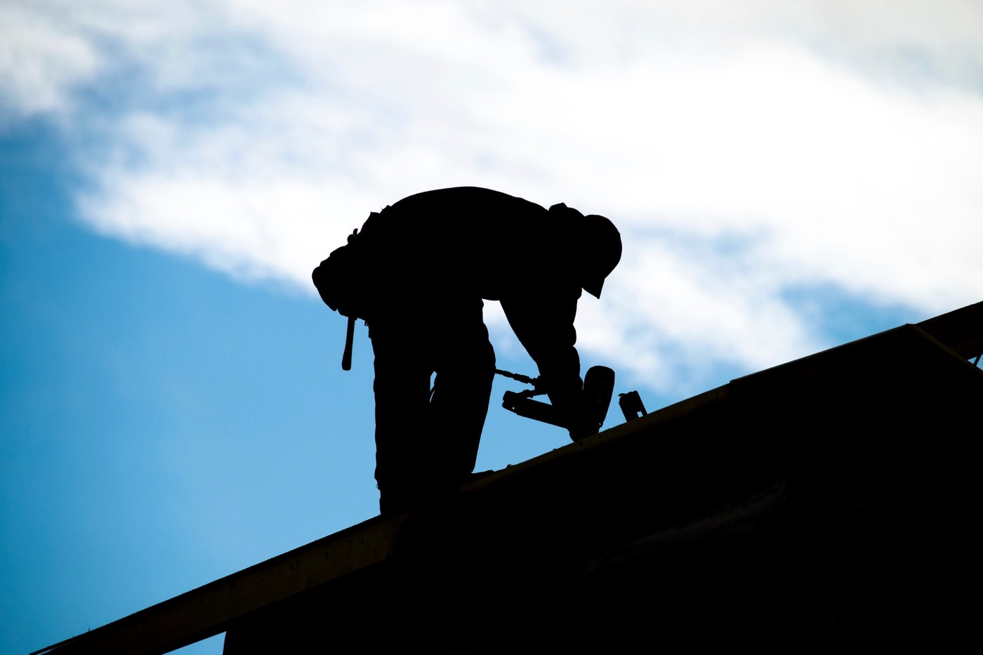 Silhouette eines Dachdeckers bei der Arbeit auf einem Dach vor blauem Himmel mit Wolken
